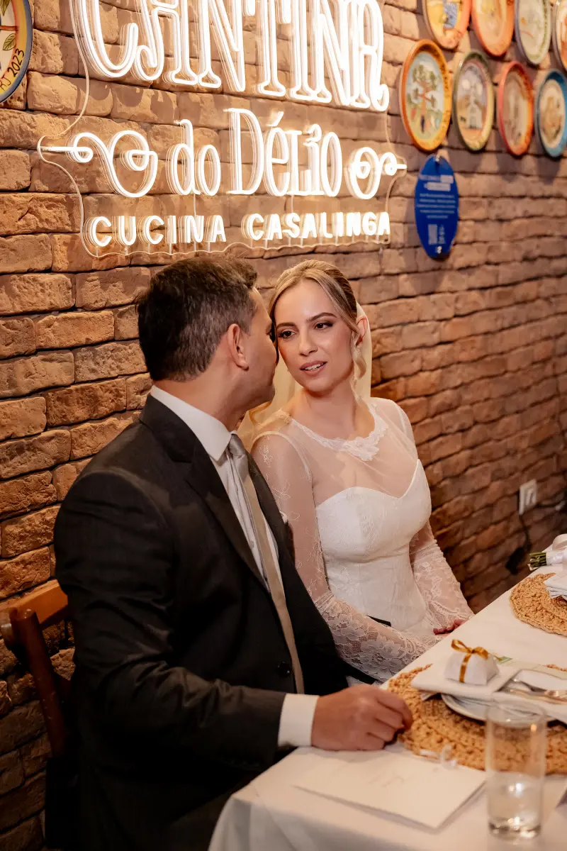 Noivos sentados à mesa sob o letreiro iluminado da Cantina do Délio